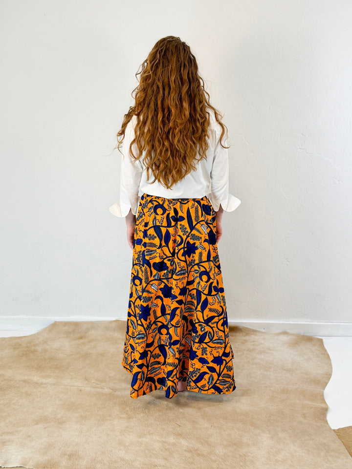 Model wearing an orange maxi skirt with bold floral pattern in dark blue. Model has paired with a long sleeve white ruffle collar shirt by Heidi Houston. Back view.