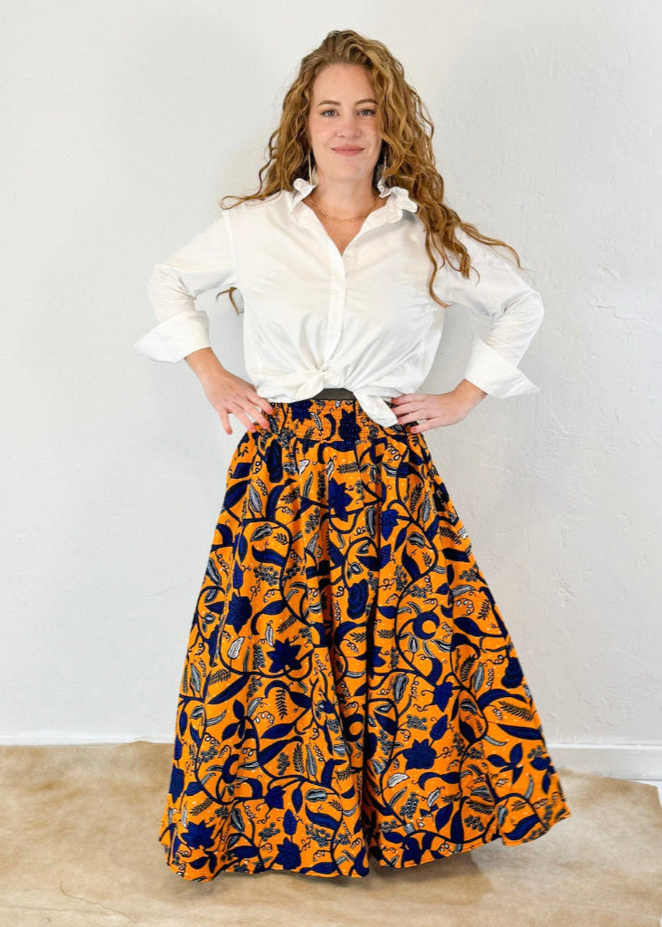 Model wearing an orange maxi skirt with bold floral pattern in dark blue. Model has paired with a long sleeve white ruffle collar shirt by Heidi Houston. Front view.